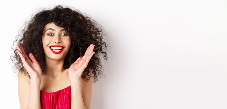 Close-up of fashionable woman with curly hair and red dress, clap hands smiling happy, applause you, standing on white background.