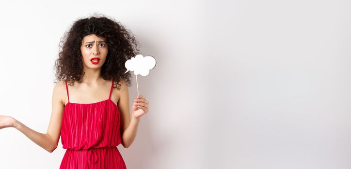 Confused young woman in red dress, frowning and looking upset, holding comment cloud stick, standing against white background.