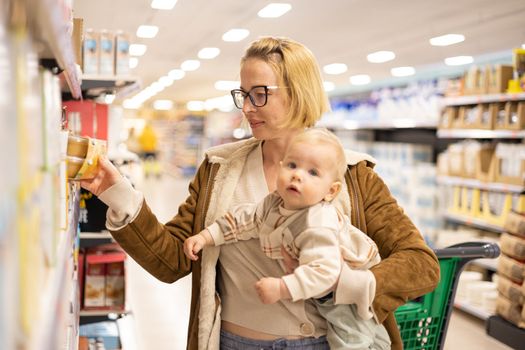 Caucasian mother shopping with her infant baby boy child choosing products in department of supermarket grocery store