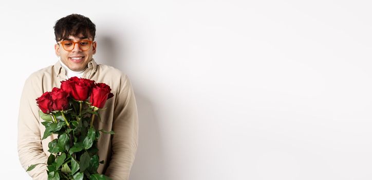 Cheerful gay man receive red roses from boyfriend, looking excited and smiling, having romantic date on Valentines day with lover, standing over white background.