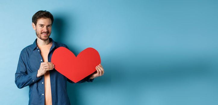 Happy man showing valentines heart and smiling, make romantic gift on lovers day, standing over blue background.