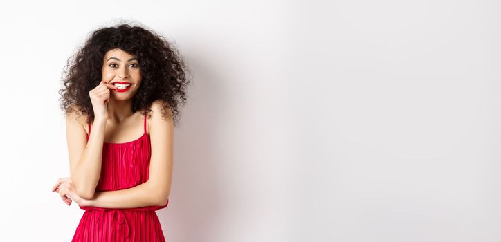 Intrigued young woman with curly hair, wearing elegant red dress and lipstick, biting fingernails and looking interested, standing over white background.