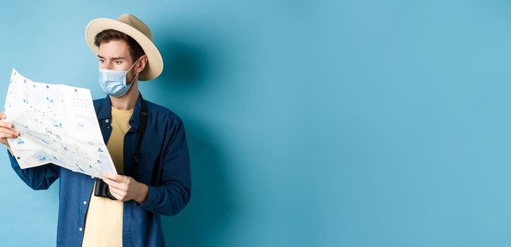 Covid-19, pandemic and travel concept. Handsome young guy in face mask and summer hat looking at tourist map, travelling on vacation, standing on blue background.