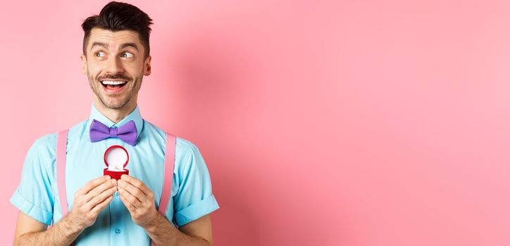 Valentines day. Romantic handsome man looking dreamy and smiling, showing engagement ring for his lover, standing happy over pink background.