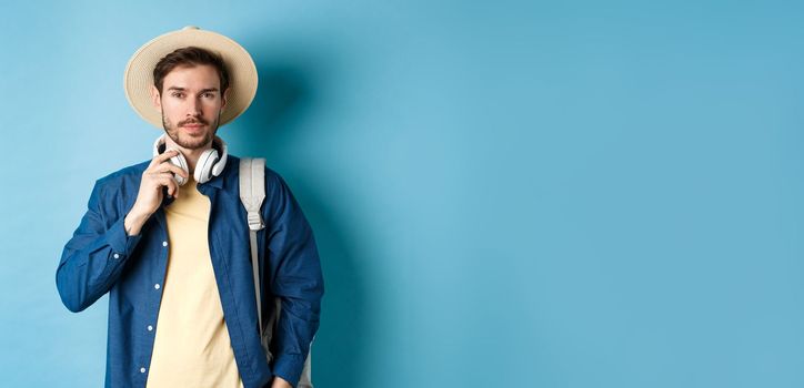 Handsome guy travelling with headphone, wearing straw hat and smiling, standing with backpack on blue background.