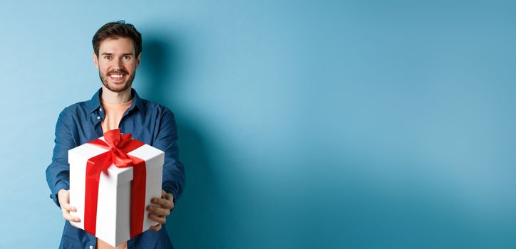 Valentines day. Handsome smiling man extending hands with gift box, wishing happy holiday. Guy making surprise present and looking cheerful, standing over blue background.