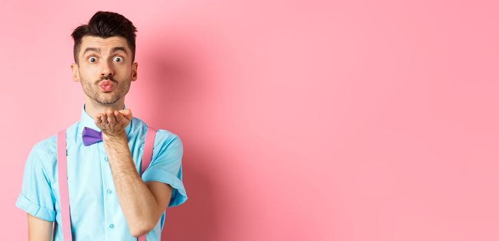 Cute boy sending air kiss at camera and saying I love you on Valentines day, standing over romantic pink background in bow-tie and shirt.