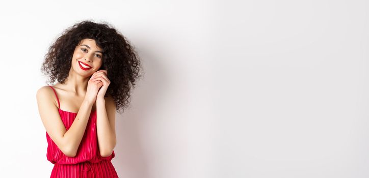 Lovely young woman with curly hairstyle, wearing red dress, looking at silly and tender thing, admire something, standing over white background.