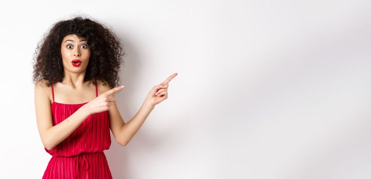 Surprised woman in red dress and makeup pointing fingers right, showing logo and look intrigued, standing on white background.