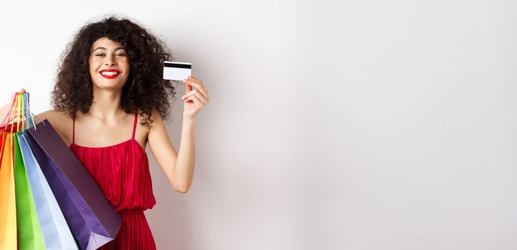 Happy elegant woman in red dress, showing shopping bags and plastic credit card, smiling pleased, standing on white background.