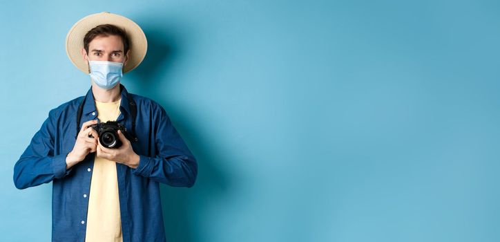 Covid-19, pandemic and travel concept. Happy guy on vacation taking pictures with camera, wearing medical mask and summer hat, blue background.