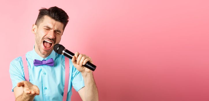 Party and festive events concept. Passionate singer holding microphone and pointing hand at you, singing about love, standing on pink background.