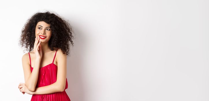 Dreamy elegant girl with curly hair, wearing red dress and makeup, looking left and smiling at logo, standing on white background.