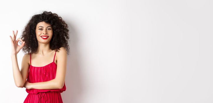 Elegant woman in romantic red dress and makeup, showing okay sign and smiling, recommending good thing, approve and agree, standing satisfied on white background.