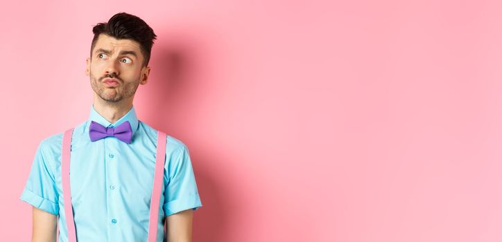 Pensive young man in romantic outfit, looking away and thinking, standing thoughtful on pink background in fancy bow-tie and shirt.
