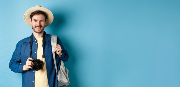 Cheerful handsome guy going on vacation, wearing summer hat and holding backpack with camera for photos, smiling excited of holiday, standing on blue background.