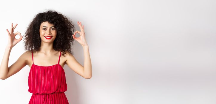 Beautiful woman in red dress, smiling and showing okay gestures, like something good, praising good job, standing over white background.