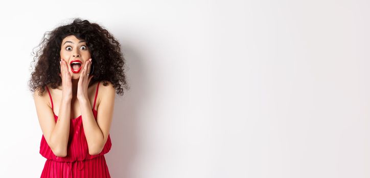 Image of elegant woman scream surprised, staring at camera, see promo offer and shouting of joy, standing in dress over white background.