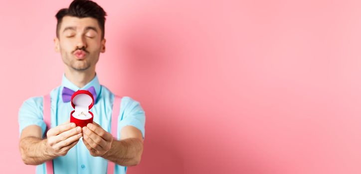 Valentines day. Silly boyfriend in bow-tie pucker lips, close eyes and waiting for kiss after making proposal, showing golden engagement ring in small red box, standing on pink background.