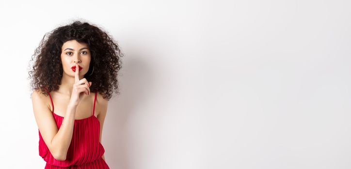 Beautiful woman with curly hair, wearing red dress, hushing at camera, telling secret, shushing at you, standing over white background.