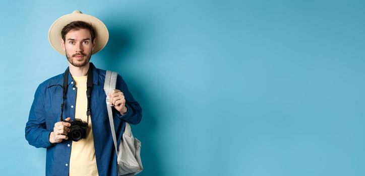 Handsome young tourist in summer hat, backpacking and travelling, holding photo camera, standing on blue background.