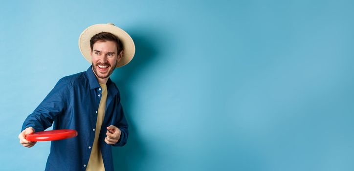 Happy man laughing and playing frisbee, throwing at friend and smiling, standing in summer hat on blue background.