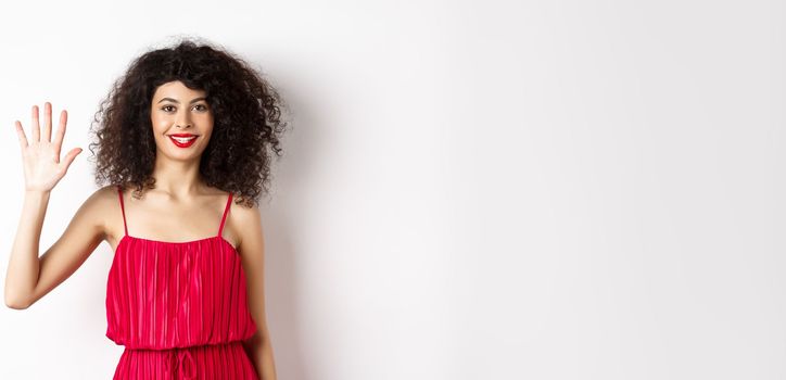 Cheerful young woman with makeup and red dress, showing five fingers and smiling, standing over white background. Copy space