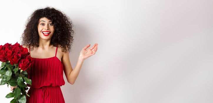 Romance and Valentines day. Woman gasping surprised and happy, receive surprise gift from lover, holding bouquet of red roses, standing on white background.