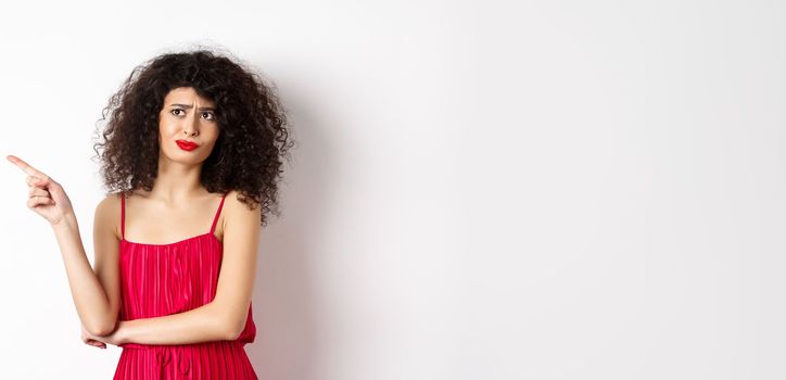 Disappointed and skeptical young woman with curly hair, wearing red dress, grimacing and pointing finger left at logo, standing over white background.