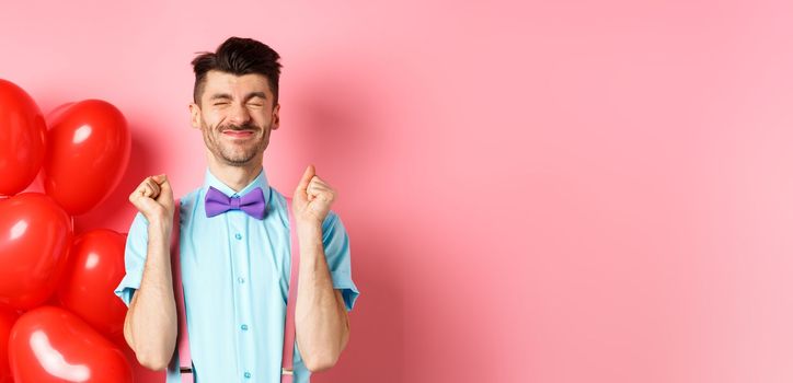 Valentines day concept. Cheerful guy jumping from excitement before romantic date, standing near big red hearts balloons and celebrating, pink background.