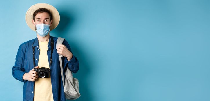 Covid-19 and summer holidays concept. Excited tourist in straw hat and medical mask looking in awe, holding camera, taking pictures on holiday during pandemic, blue background.