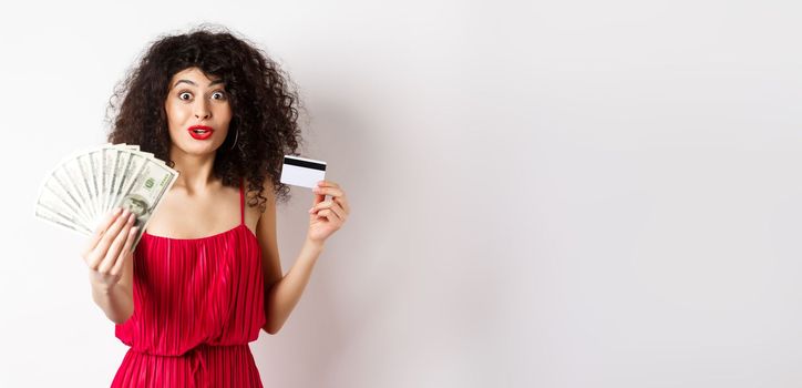 Excited woman looking with amazement and disbelief, showing dollars prize and plastic credit card, standing in red dress on white background.