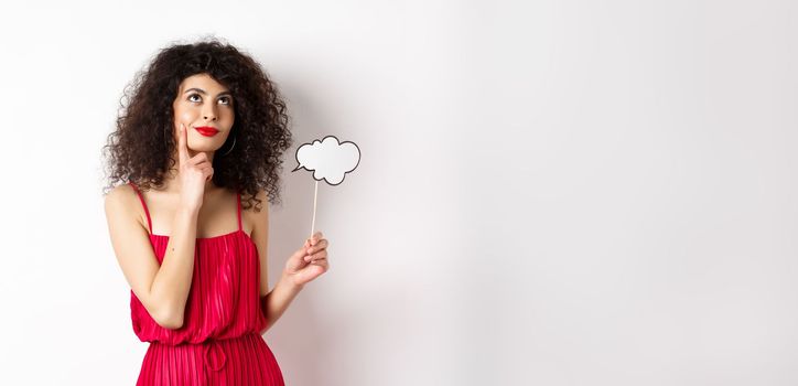 Thoughtful girl in red dress, holding small cloud and looking up with pleased smile, imaging things, standing over white background.