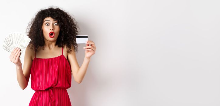 Impressed lady with curly hair and trendy red dress, saying wow, showing plastic credit card with cash, standing on white background.