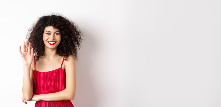 Cheerful elegant woman saying hello, waving hand and smiling at camera, greeting someone, standing in red dress on white background.