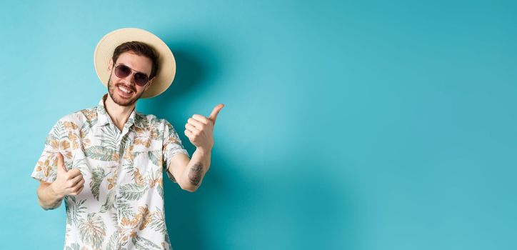 Cheerful tourist having fun summer holidays, showing thumbs up and smiling, standing in hawaiian shirt and sunglasses on blue background.