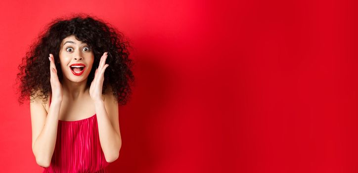 Close-up of surprised beautiful woman, screaming of amazement and looking at promo offer, standing in red dress and lipstick, studio background.