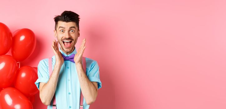 Love and romance concept. Happy man screaming from fantastic news, shouting wow and smiling amused, checking out special offer on Valentines, standing near red hearts balloons.