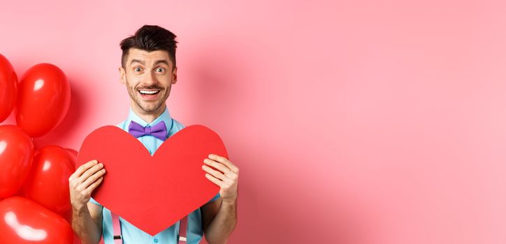 Valentines day concept. Cute young man in bow-tie showing big red heart postcard and say love you, smiling happy at camera, standing on romantic pink background.