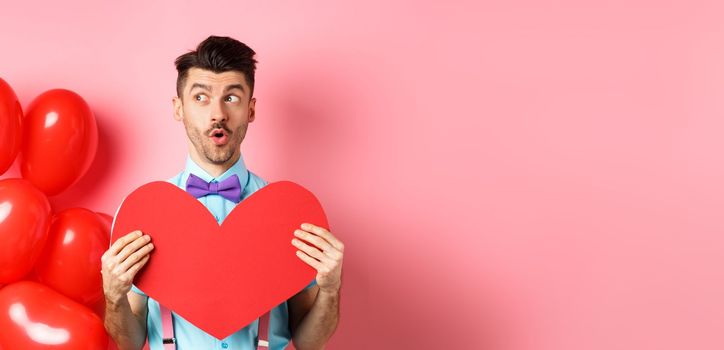 Valentines day concept. Cute young man looking left amused, showing red heart cutout and standing near balloons, pink background.