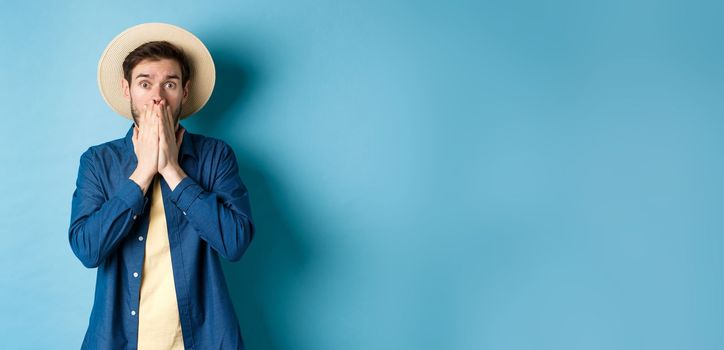 Shocked and alarmed tourist gasping, covering mouth with hands and looking startled at camera, standing on blue background.