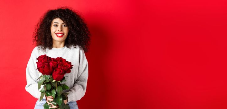 Romantic woman with curly hairstyle receive bouquet of red roses on Valentines day, looking tender at lover, standing on studio background.