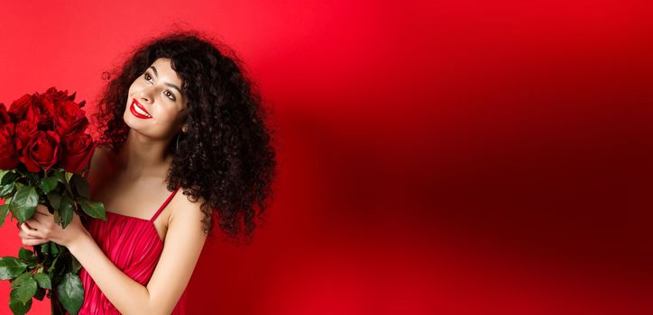 Happy beautiful woman in dress, holding flowers and smiling romantic, looking aside at logo, standing against red background.