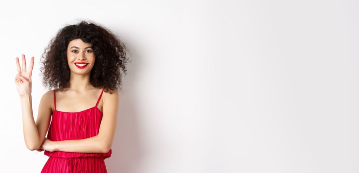 Beautiful lady with curly hair, wearing elegant red dress and showing number three, making order, standing over white background.