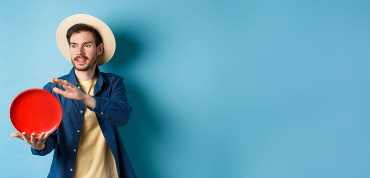 Happy smiling guy throwing frisbee, looking aside at friend, standing on blue background in straw hat.