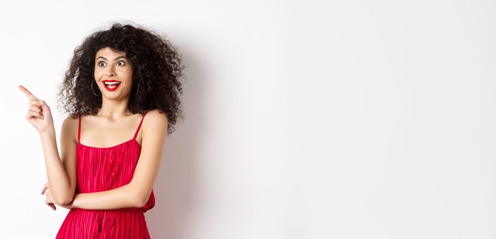 Surprised female model with curly hair and makeup, pointing finger and looking left with amused smile, standing in red dress over white background.