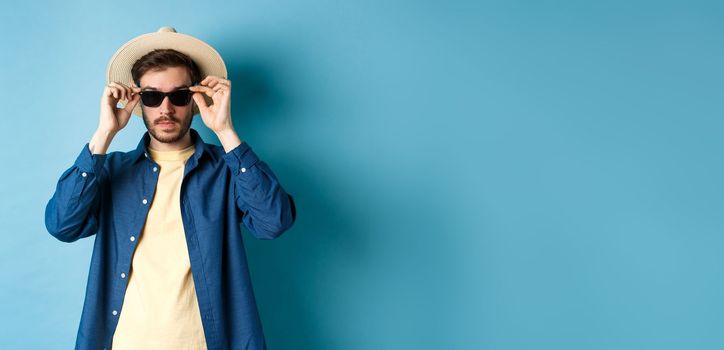 Handsome tourist in straw hat put on sunglasses on summer vacation, standing on blue background.