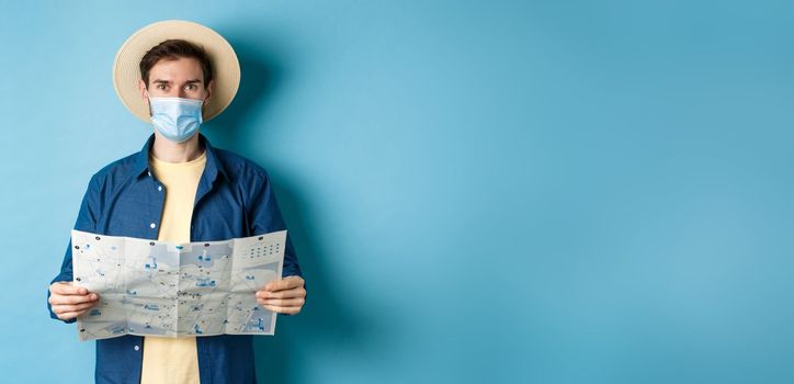 Covid-19, pandemic and travel concept. Happy tourist on summer vacation wearing medical mask and straw hat, holding road map, standing on blue background.