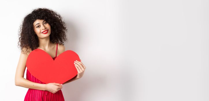 Romantic tender woman with curly hair, hugging big red heart and smiling, look with love, standing against white background.