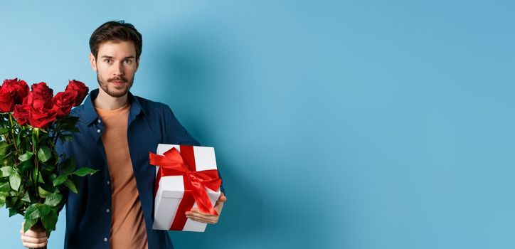 Romance and valentines day. Man presenting bouquet of red roses to lover. Boyfriend bring flowers and gift on romantic date, standing over blue background.